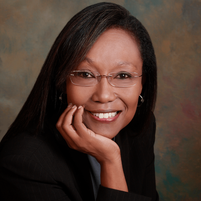 Smiling woman in a business suit, resting her chin on her hand, against a softly colored background. Represents professionalism and confidence.