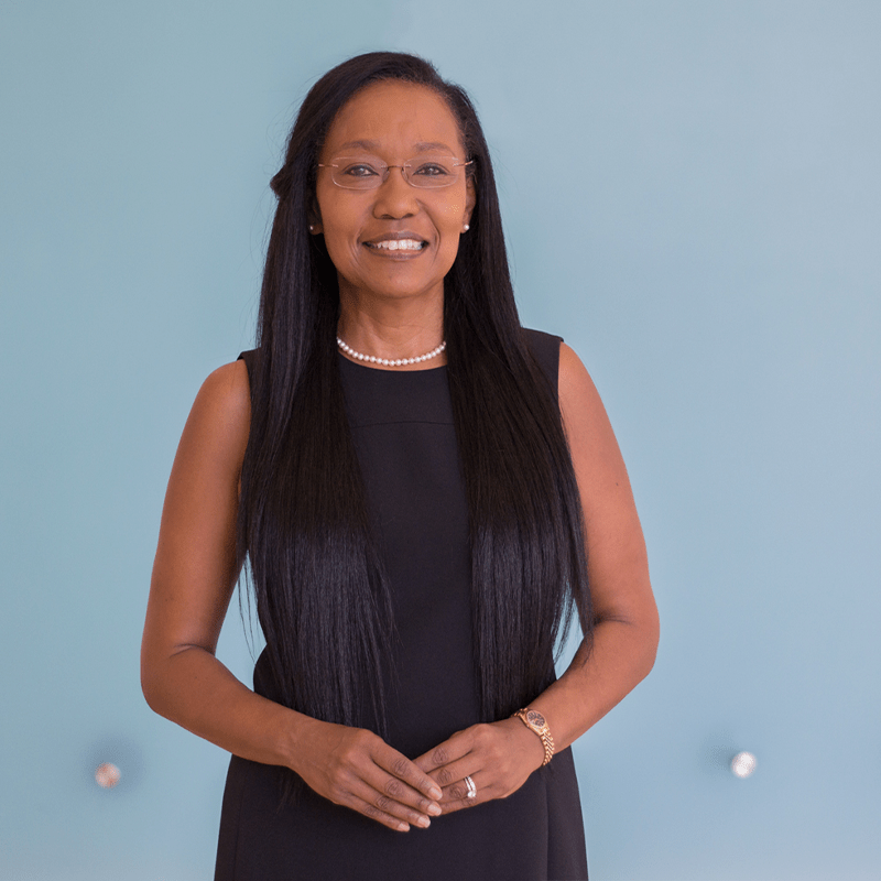 A confident woman with long hair, wearing a black dress and pearl necklace, stands against a light blue background, smiling warmly.
